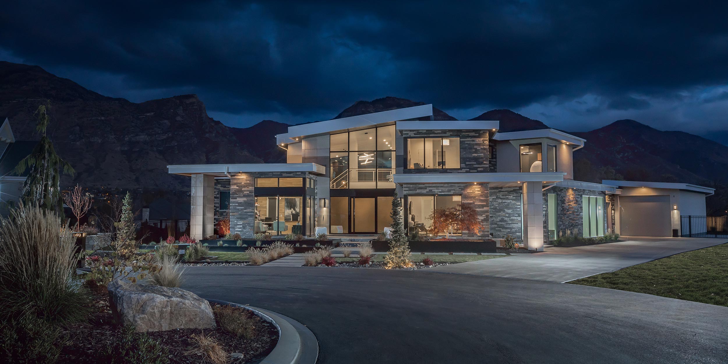 An evening view of a luxurious, contemporary home with dramatic lighting set against a mountainous backdrop.