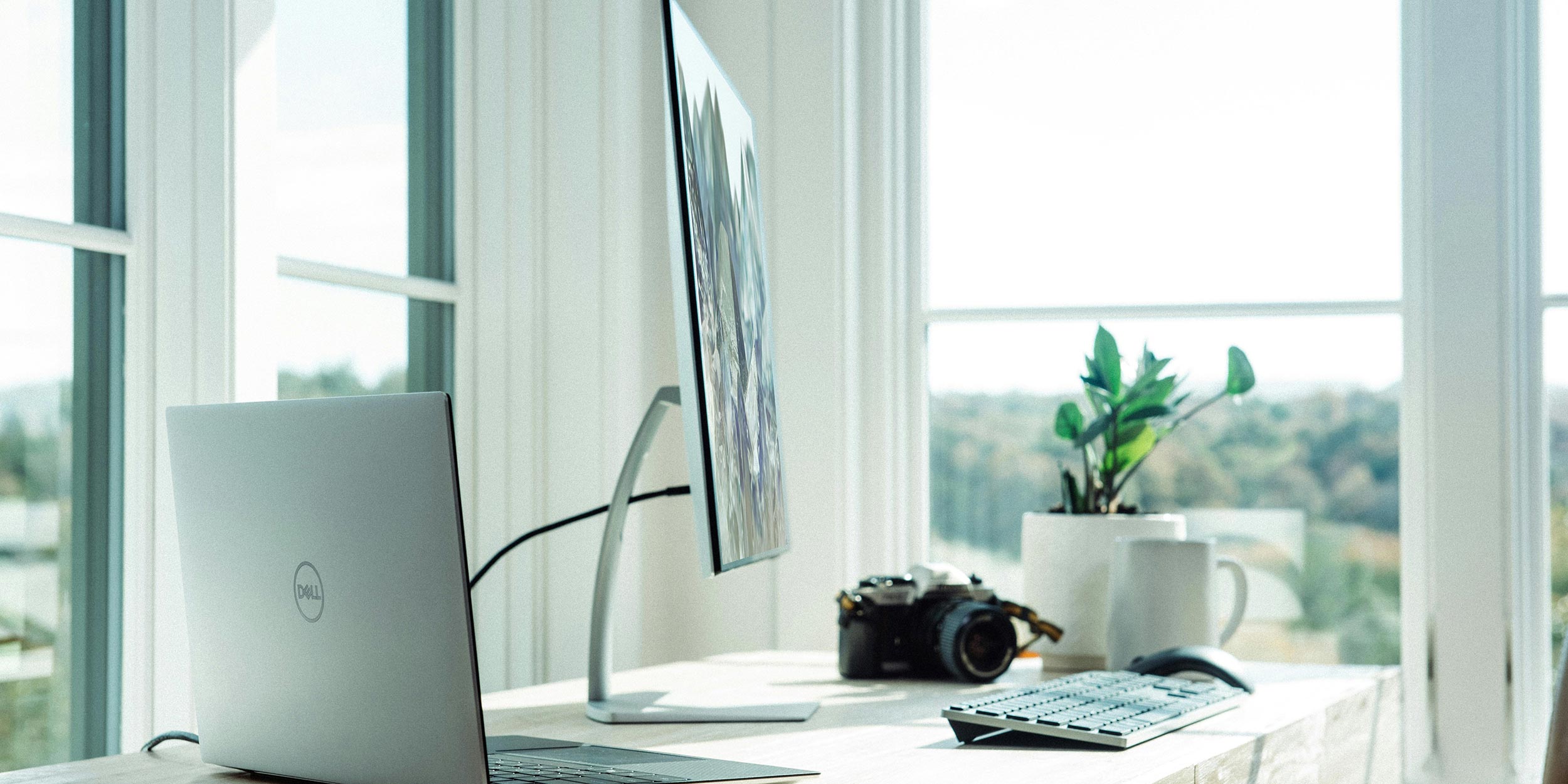 A bright workspace with a laptop, monitor, and a camera placed on a desk near large windows.